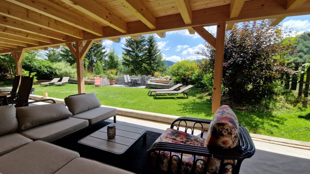 Überdachte Lounge im Garten des Kräuterhäusl mit Sofa, Liegestühlen und Blick auf die Feuerstelle